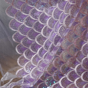 Op de afbeelding: Close-up van een stof met een zeemeerminstaartpatroon. De stof heeft een herhalend ontwerp van overlappende, halfronde schubben in tinten paars en lavendel, versierd met pailletten. De achtergrond is een doorschijnend, wit gaas.