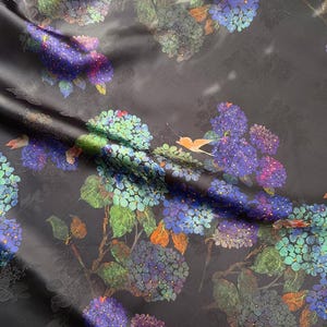 May include: Dark fabric with a floral design of blue, purple, and green hydrangeas. The pattern includes leaves and orange accents, creating a botanical theme. The fabric has a subtle sheen.