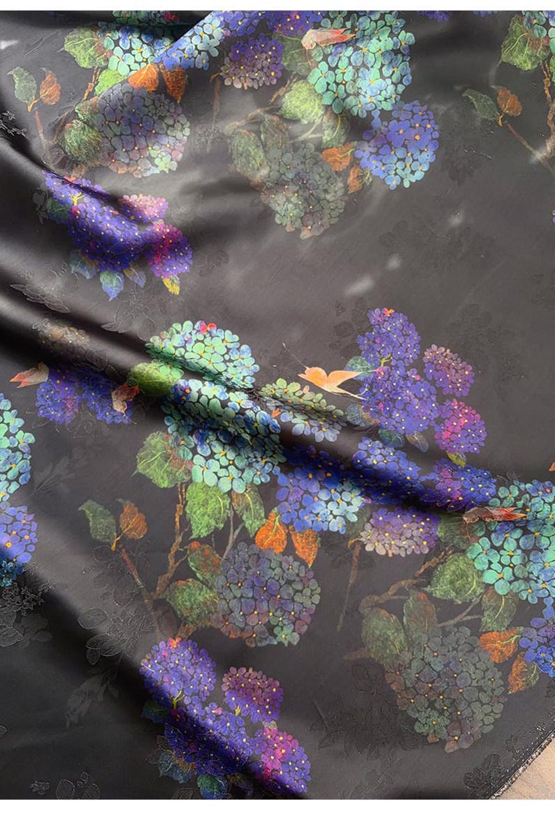 May include: Dark fabric with a floral design of blue, purple, and green hydrangeas. The pattern includes leaves and orange accents, creating a botanical theme. The fabric has a subtle sheen.