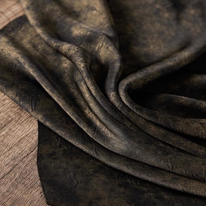 Puede incluir: Una pieza de tela con un color base negro oscuro y un acabado dorado metálico desgastado aplicado en la superficie. El material está drapeado en pliegues suaves, resaltando el contraste entre los tonos negros profundos y el oro brillante.