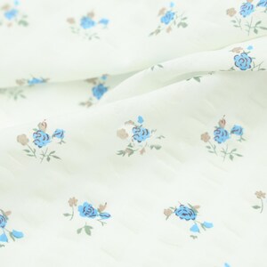 May include: White fabric with a delicate floral pattern. Small blue flowers with brown and green accents are scattered across the surface. The fabric has a subtle texture and is slightly wrinkled, suggesting it is soft and pliable.