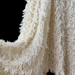 May include: A cream-coloured fabric featuring a dense, shaggy texture with long, frayed, thread-like fibres covering the entire surface. The material has a layered, fringe-like appearance with a consistent off-white hue throughout the textile.