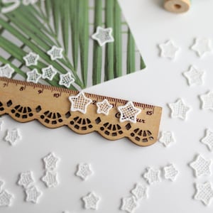 May include: White lace star-shaped embellishments scattered on a white surface with a wooden ruler and green foliage in the background.