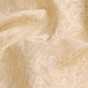 May include: Close-up of a cream-colored fabric with an embroidered floral pattern. The fabric has a delicate, sheer quality, with subtle variations in texture and tone. The embroidery adds a touch of elegance to the textile.