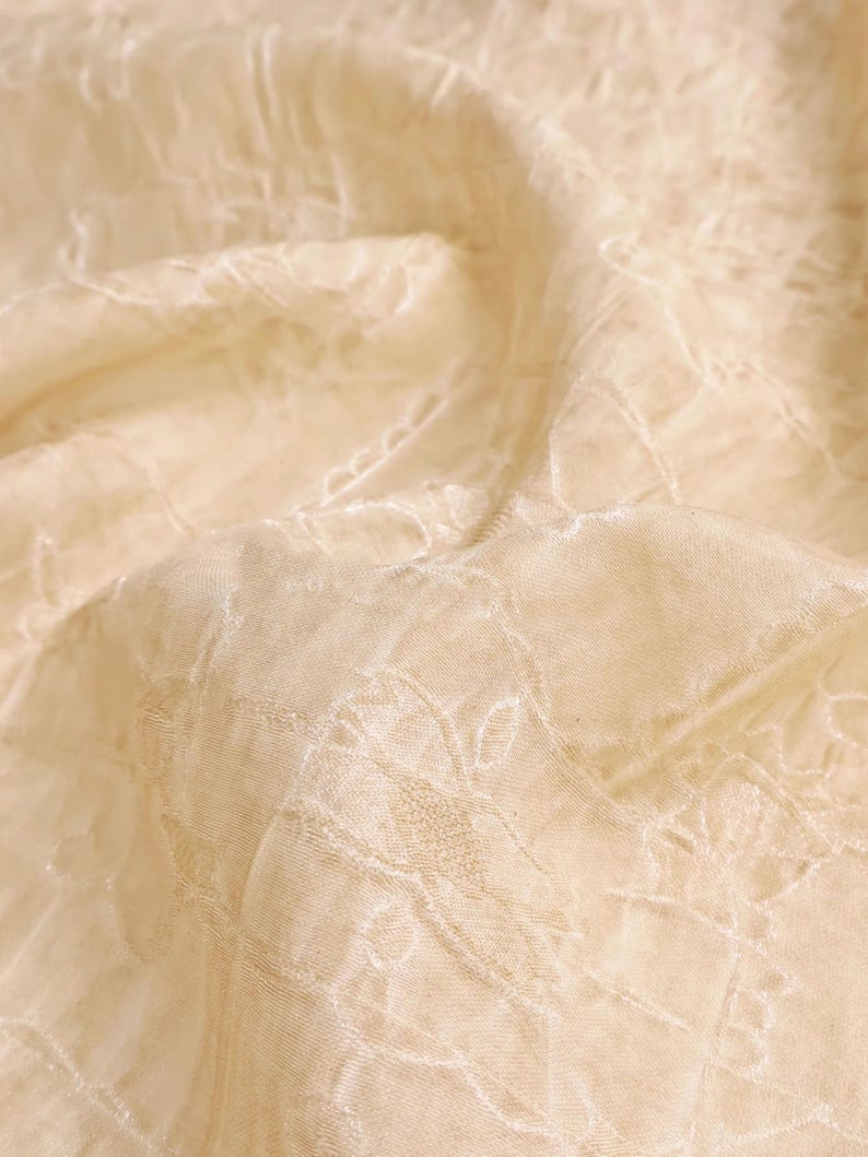 May include: Close-up of a cream-colored fabric with an embroidered floral pattern. The fabric has a delicate, sheer quality, with subtle variations in texture and tone. The embroidery adds a touch of elegance to the textile.