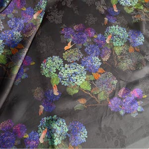 May include: Dark fabric with a floral pattern featuring blue, purple, and green hydrangea blooms. The design includes orange butterfly accents and subtle grey floral outlines on a black background. Suitable for craft projects.