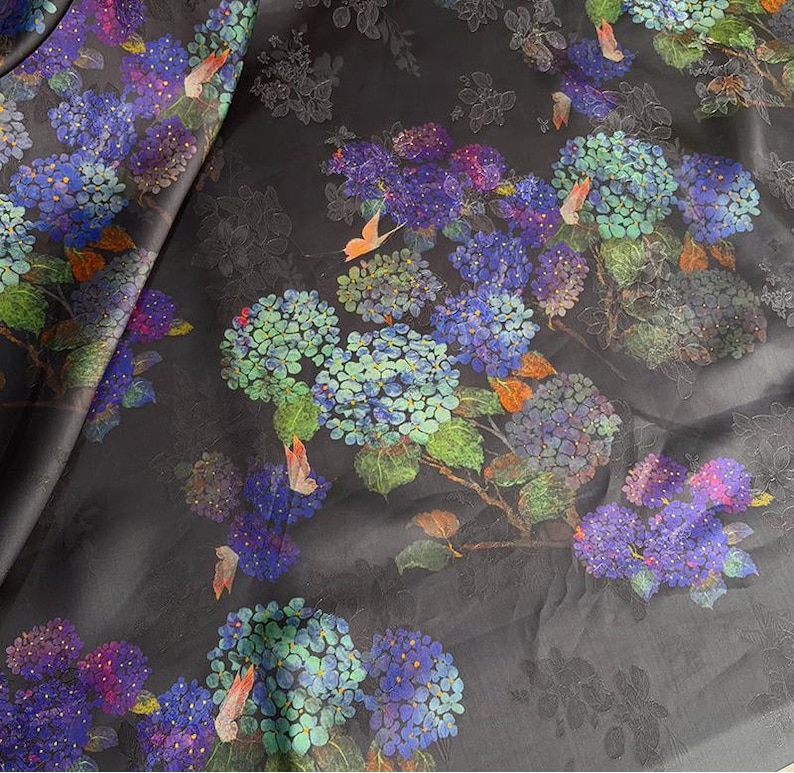 May include: Dark fabric with a floral pattern featuring blue, purple, and green hydrangea blooms. The design includes orange butterfly accents and subtle grey floral outlines on a black background. Suitable for craft projects.