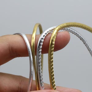 May include: Close-up of four metal bangles in silver and gold tones. The bangles have varying textures, with some featuring a hammered or textured surface. The background is a solid, neutral gray.
