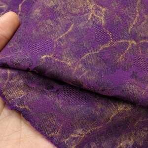 May include: A deep purple fabric featuring a woven metallic gold pattern. The textile displays a complex design of interconnected lines and textured geometric shapes that create a shimmering effect across the surface of the folded material.