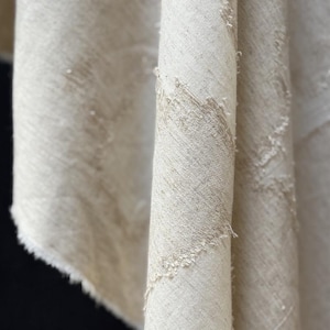 May include: Close-up of a rolled, off-white fabric with a textured, diagonal weave. The fabric has a raw, frayed edge and a subtle, natural color variation. The material appears to be linen or a similar natural fiber.