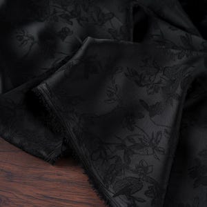 May include: Black fabric with a floral pattern. The fabric is folded, revealing the texture and design. The floral pattern is subtly visible against the black background. The fabric appears to be a luxurious material, possibly silk or satin.
