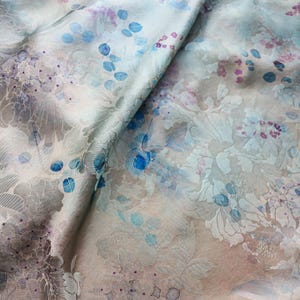 Peut inclure: Gros plan sur un tissu à motifs floraux dans les tons de bleu, violet et gris. Le tissu a un aspect soyeux et une apparence délicate et superposée. Le motif présente des motifs floraux et de feuilles détaillés.