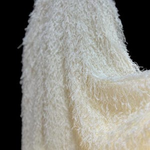 May include: Close-up of a white fluffy fabric with a soft, fuzzy texture. The fabric is draped and folded, creating a sense of movement and texture.