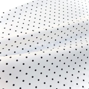 Op de afbeelding: Witte stof met een patroon van gelijkmatig verdeelde zwarte stippen. De stof is licht gekreukt, waardoor zachte schaduwen en highlights ontstaan. De stippen zijn consistent in grootte en vorm, wat een klassiek ontwerp biedt.