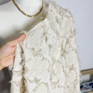 Puede incluir: Tejido blanquecino con un diseño texturizado y con flecos. La tela está drapeada sobre un maniquí, y se ve un collar de cadena de oro y plata. La textura de la tela se destaca por los bordes deshilachados y los patrones en relieve.