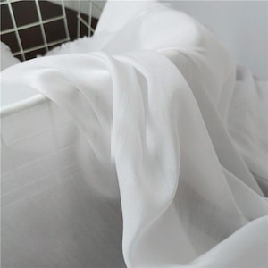 May include: Close-up of white fabric draped over a white metal basket. The fabric is sheer and flowing, with soft folds and highlights. The texture appears smooth and delicate, suggesting a lightweight material.