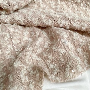 May include: Close-up of a textured fabric with a woven pattern. The fabric is a blend of light pink and off-white, with small, raised, nubby details and scattered sequins. The material is folded, revealing a glimpse of a white surface underneath.