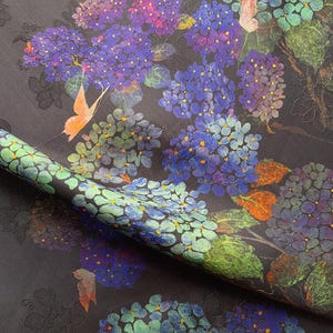 May include: Dark fabric with a floral pattern featuring blue, purple, and green hydrangeas and orange butterflies. The fabric has a textured surface and a rolled edge, showcasing the detailed design.