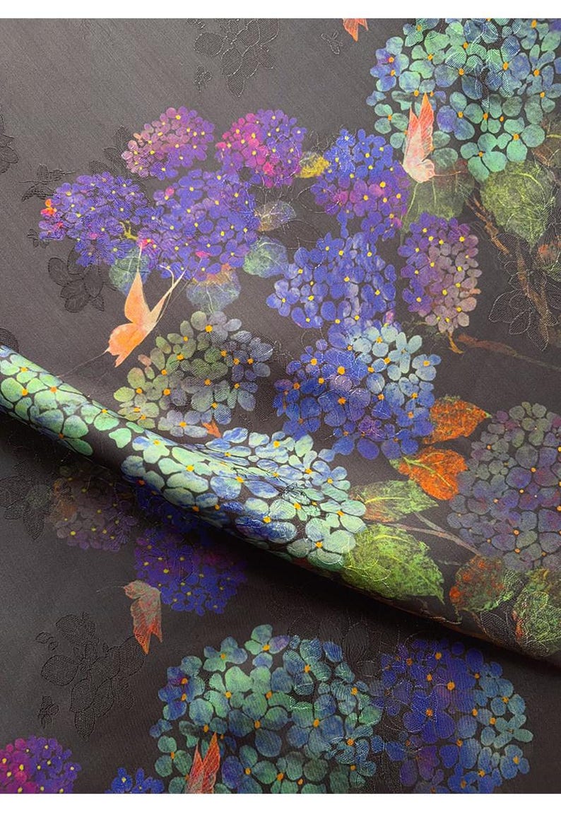 May include: Dark fabric with a floral pattern featuring blue, purple, and green hydrangeas and orange butterflies. The fabric has a textured surface and a rolled edge, showcasing the detailed design.
