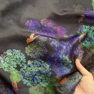 May include: Dark fabric with a floral design. The pattern features clusters of blue, purple, and green flowers. The fabric has a subtle sheen and a dark background, with hints of orange butterflies. The design is detailed and appears to be a silk or similar material.