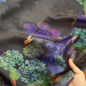 May include: Dark fabric with a floral design of purple, blue, and green flower clusters. The background is dark grey with subtle embossed floral details. The fabric is held, showing its texture and drape.