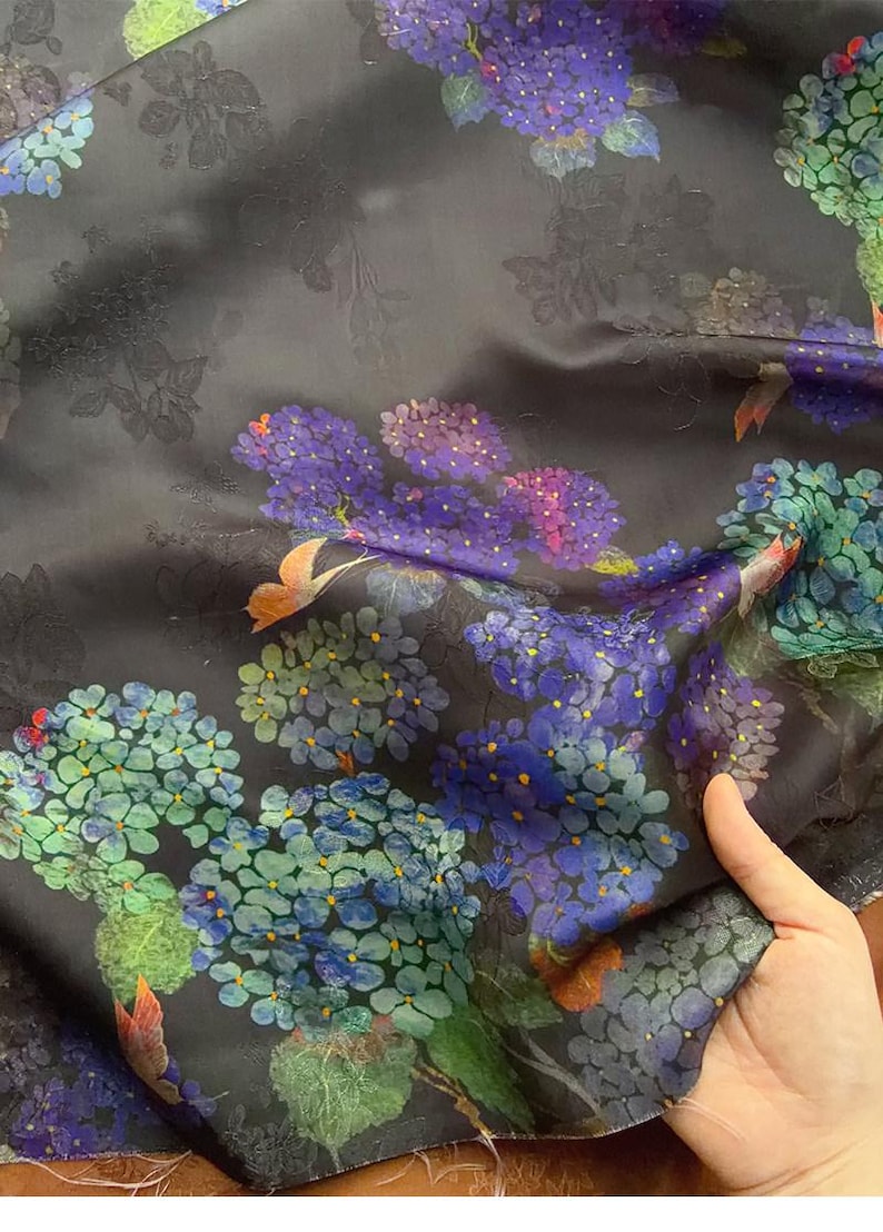 May include: Dark fabric with a floral design of purple, blue, and green flower clusters. The background is dark grey with subtle embossed floral details. The fabric is held, showing its texture and drape.