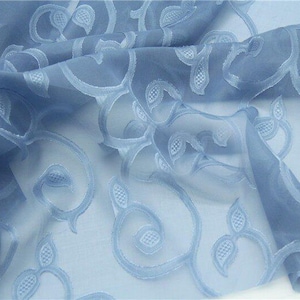 Puede incluir: Tela azul claro transparente con un bordado floral. La tela está doblada, mostrando el intrincado diseño de vides arremolinadas y formas de hojas. El material delicado tiene una apariencia suave y fluida, adecuado para cortinas o ropa.