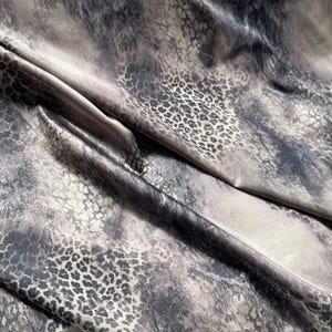 Pode incluir: Close-up de um tecido com um design de estampa de leopardo. O tecido tem uma mistura de tons cinza, preto e bege, com uma textura brilhante e lisa. As manchas de leopardo estão agrupadas em algumas áreas e espalhadas em outras, criando um padrão dinâmico.