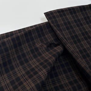 May include: Close-up of a dark fabric with a brown plaid pattern. The fabric is folded, showing the texture and the interplay of light and shadow. The plaid design consists of intersecting lines creating a grid-like appearance.
