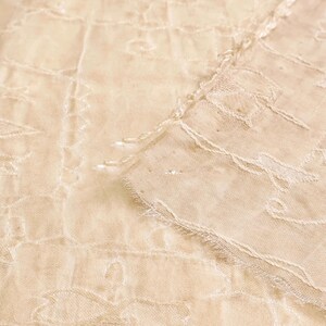 May include: Close-up of a cream-colored, sheer fabric with an intricate floral pattern. The fabric has a delicate, textured appearance with visible stitching and a frayed edge, suggesting a vintage or handmade textile.
