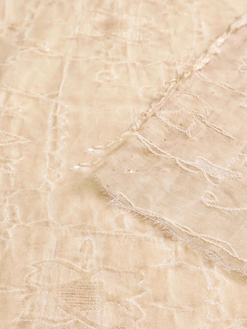 May include: Close-up of a cream-colored, sheer fabric with an intricate floral pattern. The fabric has a delicate, textured appearance with visible stitching and a frayed edge, suggesting a vintage or handmade textile.
