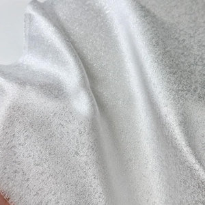 May include: A piece of fabric with a shimmering silver and white finish. The material features a fine, glittery texture that catches the light, creating subtle highlights and deep shadows across the folds of the draped, reflective textile surface.