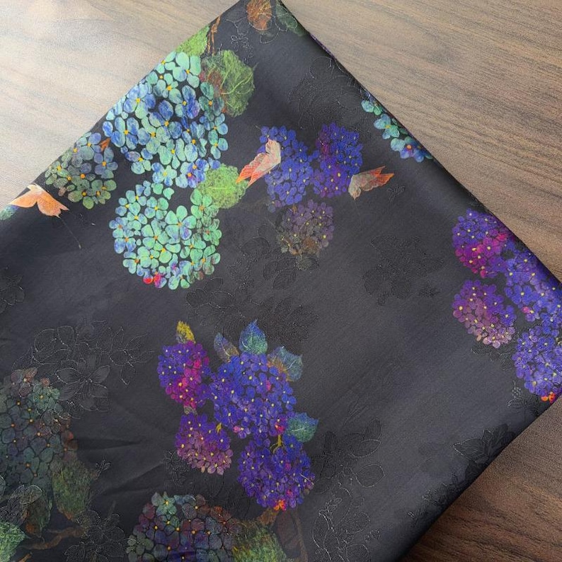 May include: Dark fabric with a floral design of blue, purple, and green hydrangea flowers. The fabric has a slight sheen and a black background, creating a contrast with the colorful flowers. The fabric is folded.