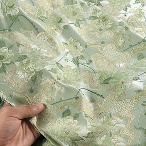 Puede incluir: Primer plano de una tela verde claro con un intrincado diseño floral y de criaturas míticas. El patrón presenta flores y criaturas míticas bordadas en oro y verde. La tela tiene un brillo satinado.