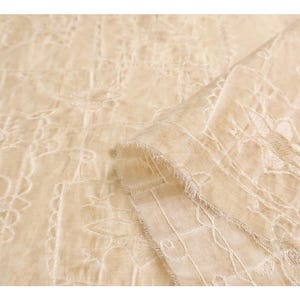 May include: Close-up of a beige fabric with intricate white embroidery. The fabric is folded, revealing a layered texture and a delicate, fringed edge. The embroidery features floral and geometric patterns.
