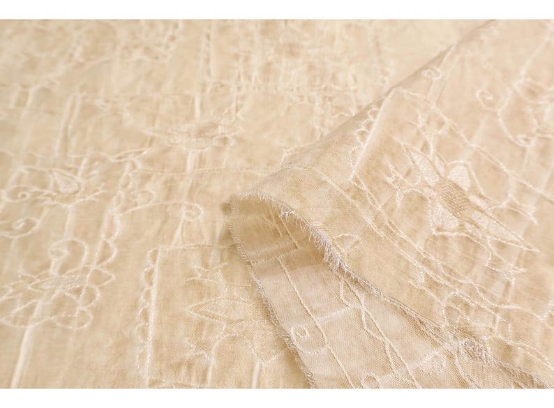 May include: Close-up of a beige fabric with intricate white embroidery. The fabric is folded, revealing a layered texture and a delicate, fringed edge. The embroidery features floral and geometric patterns.