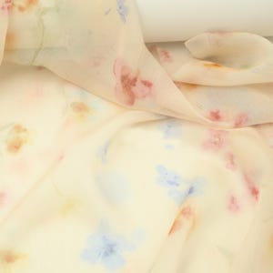 May include: A close-up of a cream-coloured fabric with a delicate floral pattern. The design features watercolour-style flowers in shades of pink, blue, and yellow. The fabric is draped, showing its soft texture and subtle sheen, ideal for apparel or home decor.