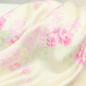 Puede incluir: Tela color crema con un estampado floral de peonías rosas y hojas verdes. La tela está drapeada, mostrando la textura suave y los pliegues del material. El diseño floral está distribuido uniformemente por la tela.