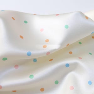 Op de afbeelding: Close-up van een crèmekleurige stof met een satijnglans. De stof is versierd met verspreide stippen in pastelkleuren: oranje, groen, roze en blauw. De stof is zachtjes gevouwen, waardoor zachte schaduwen en highlights ontstaan.