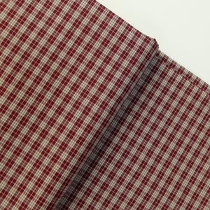 Op de afbeelding: Een gevouwen stuk stof met een rood en gebroken wit geruit patroon. De stof is over zichzelf gevouwen en toont de textuur en het weefsel van het materiaal. Het patroon is een klassiek ruitjesontwerp, geschikt voor diverse naaiprojecten.