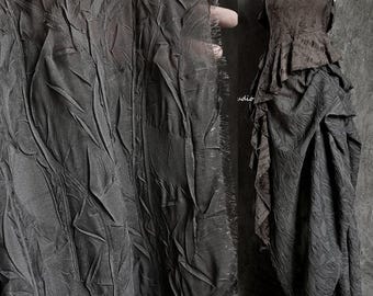 May include: Close-up of a dark gray textured fabric with a frayed edge. The fabric is draped over a mannequin wearing a dark gray dress with a ruffled hem.