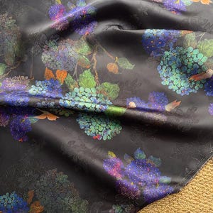 May include: Black fabric with a floral design, showcasing clusters of blue, purple, and green flowers. The pattern includes leaves and small orange bird accents. The fabric has a subtle sheen and a textured appearance.
