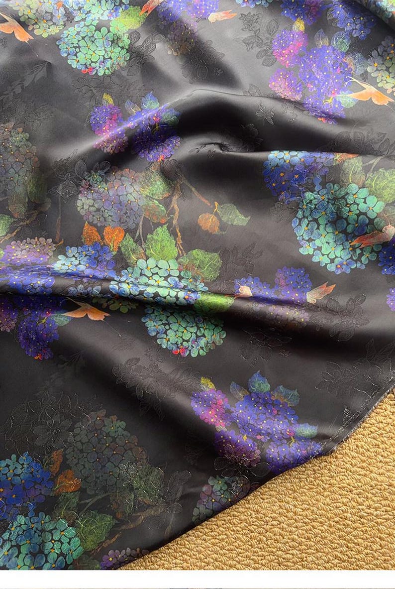 May include: Black fabric with a floral design, showcasing clusters of blue, purple, and green flowers. The pattern includes leaves and small orange bird accents. The fabric has a subtle sheen and a textured appearance.