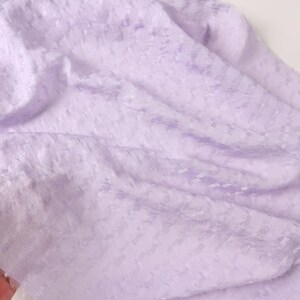 May include: A close-up shot of a soft, light purple fabric with a subtle textured pattern. The fabric is draped, creating gentle folds and shadows. The material appears to be lightweight and possibly sheer, with a delicate, almost ethereal quality.