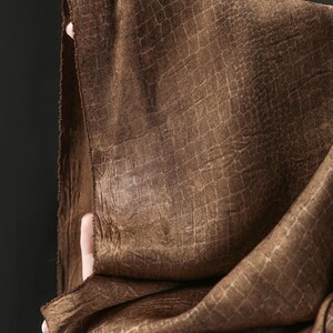 May include: A brown fabric featuring a textured, embossed pattern that resembles reptile skin. The material has a matte finish with subtle variations in tone, showing a grid-like scale design across the surface and a raw, unfinished edge.