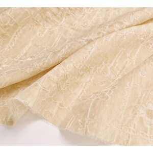 May include: Close-up of a cream-colored fabric with intricate white floral embroidery. The fabric is draped, showing the texture and detail of the design. The edges are unfinished, adding to the delicate appearance. Ideal for apparel or home decor.