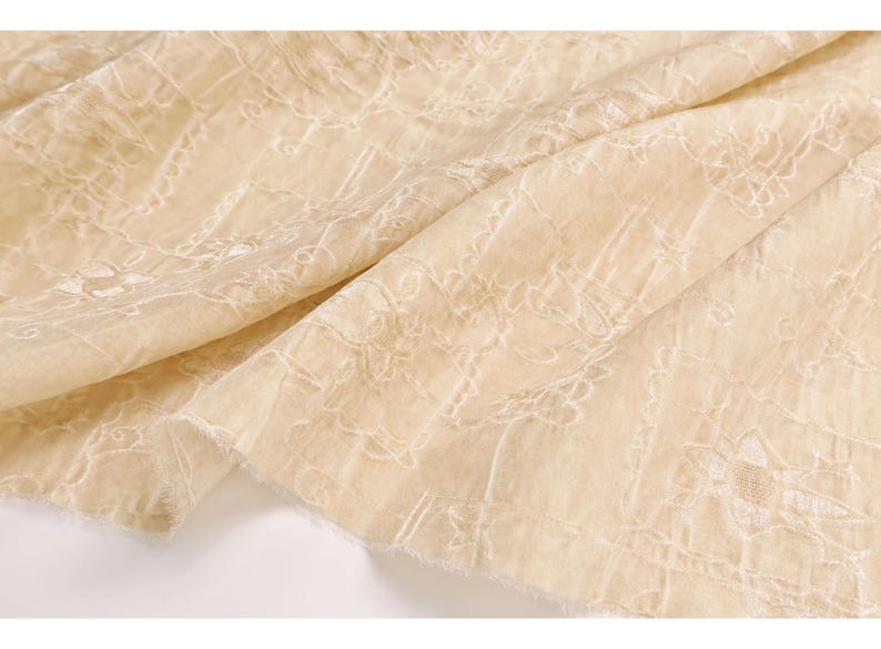 May include: Close-up of a cream-colored fabric with intricate white floral embroidery. The fabric is draped, showing the texture and detail of the design. The edges are unfinished, adding to the delicate appearance. Ideal for apparel or home decor.