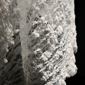 Puede incluir: Primer plano de una tela blanca texturizada con un diseño poroso en capas. La tela parece estar hecha de un material suave, con un tejido delicado y abierto. El fondo es negro, resaltando los intrincados detalles de la tela.