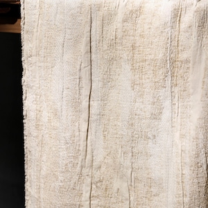May include: Close-up of a beige linen table runner with a textured, woven design. The fabric has vertical seams and a subtle, natural colour variation. The runner is draped over a wooden table, with a dark background.