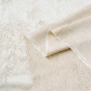 May include: A folded piece of beige fabric with a subtle, textured pattern. The fabric is layered over a larger piece of the same material, creating a sense of depth and showcasing the intricate design. The overall aesthetic is soft and neutral.